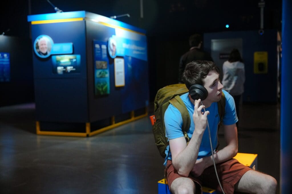 A young man with a backpack sits on a bench in the Intrepid Museum’s Mysteries from the Deep exhibition, featured by The Smithsonian, listening to audio on headphones while looking to his right. Other visitors view displays in the background.