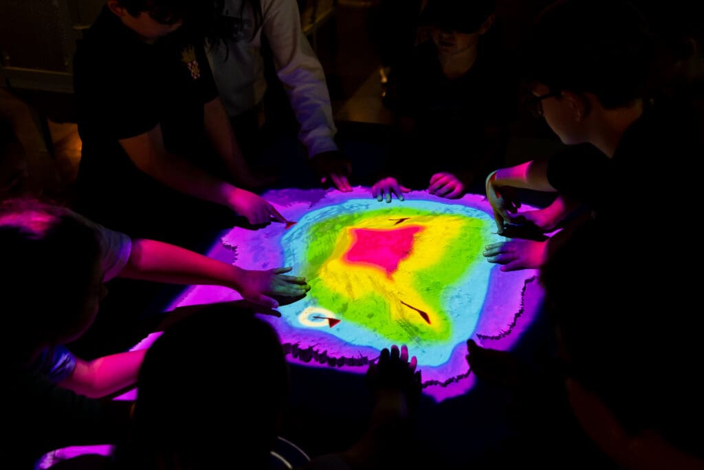 Children surround and interact with an illuminated sandbox displaying colorful topographic elevation projections, reminiscent of the mapping technology used in Finding Titanic: The Secret Mission.