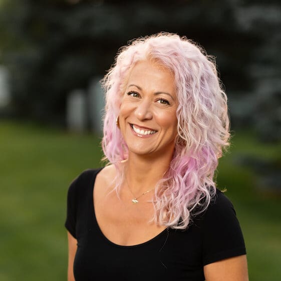 A woman with curly, light purple hair smiles outdoors, wearing a black top and a necklace. The background is blurred greenery.