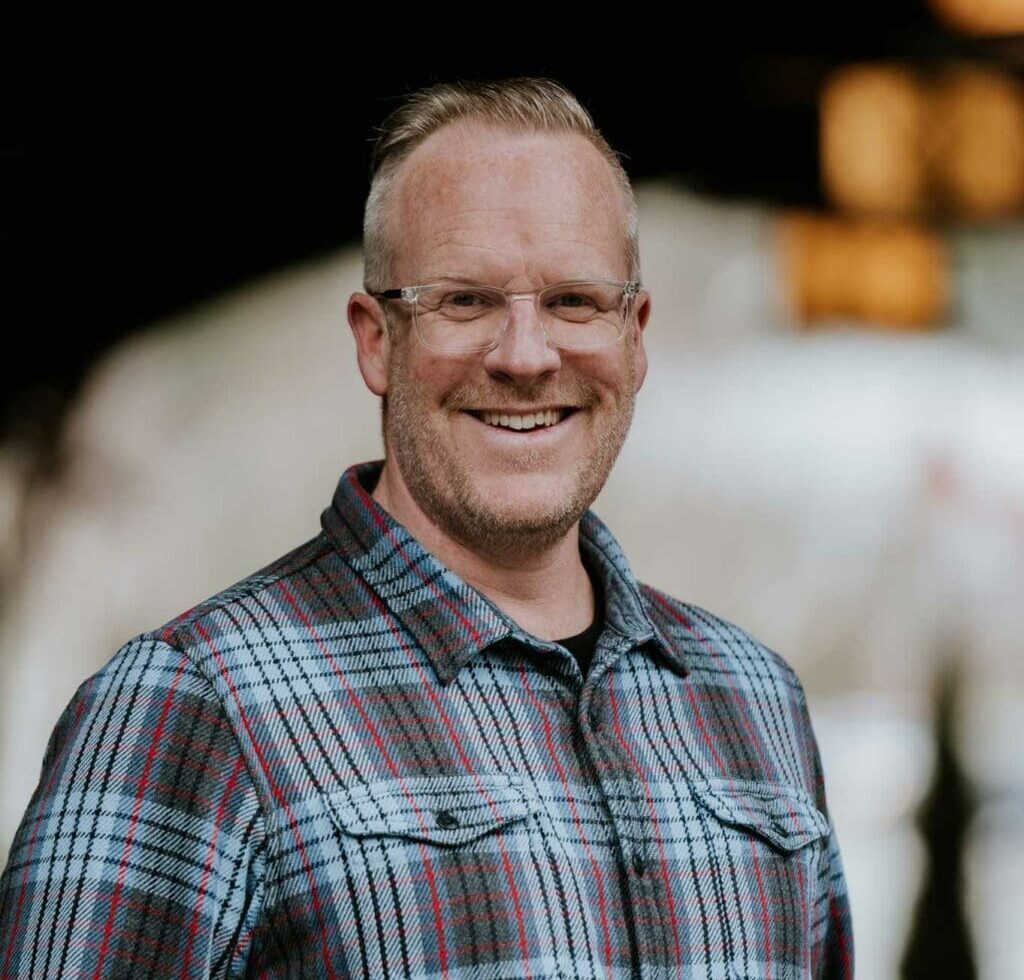 A man with short blond hair, glasses, and a beard is smiling at the camera. He is wearing a plaid shirt and standing in front of a blurred outdoor background.