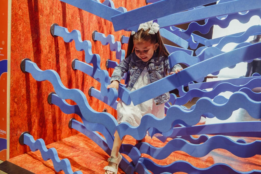 A young girl in a white dress and denim jacket steps through wavy blue foam beams in an indoor play area with red walls, capturing a moment of playful adventure in the traveling exhibition Uniquely Us.