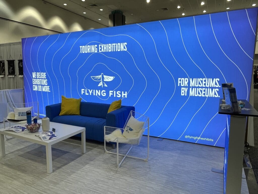 a blue wall with white text and a couch and a coffee table; the Flying Fish booth at AAM 2025