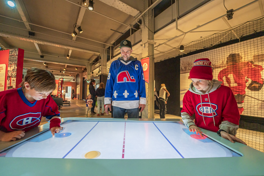 A family engages with an interactive air hockey table at the Hockey: Faster Than Ever traveling exhibition, with exhibition graphics and displays visible in the background.